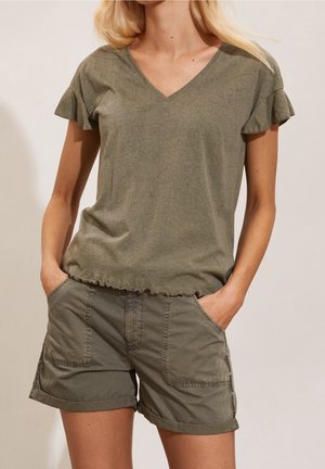 Femme portant un haut vert olive à col en V avec manches à volants et un short cargo assorti avec ourlet roulé, mains dans les poches, debout devant un fond clair.