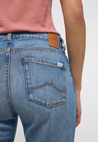 Jeans de mezclilla azul claro con un parche de marca de cuero marrón; cuentan con detalles de costura y una forma de bolsillo distintiva con un diseño de línea curva.