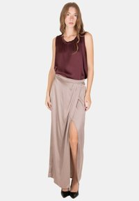 Donna che indossa un top bordeaux senza maniche e una gonna a portafoglio beige a vita alta con spacco sulla coscia, in piedi davanti a uno sfondo bianco semplice.