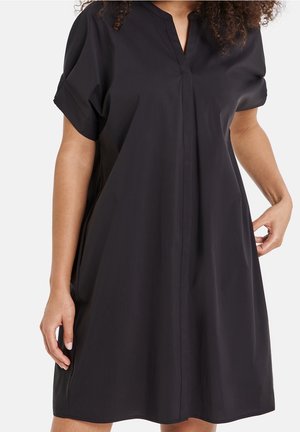 Woman wearing loose-fitting black short-sleeve dress with V-neck and hidden front seam, standing against a plain background.