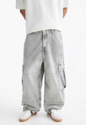 Personne portant un pantalon cargo gris ample et des baskets blanches à rayures noires, debout devant un fond blanc uni.