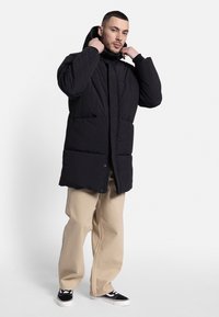 Abrigo acolchado negro con capucha, que presenta un cierre de cremallera en el frente, grandes bolsillos y una textura acolchada. Llevado sobre pantalones beige y zapatillas negras.