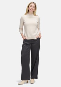 Vrouw die glimlacht, in een crèmekleurige lange mouwen top, zwarte wijde broek en crèmekleurige enkellaarzen, staande met handen in de zakken op een witte achtergrond.