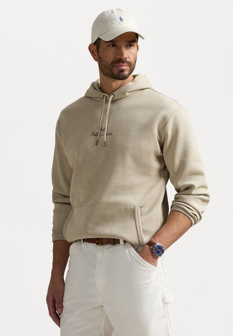 Beige hoodie med snoretræk, forlomme og broderet logo. Pairing med hvide bukser og et ur med blå skive, suppleret med en kasket.