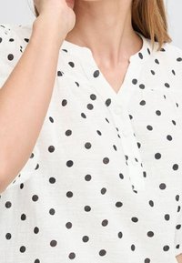 Femme portant une chemise blanche à pois noirs et boutons, main levée près du visage, montrant le cou et une partie des cheveux.