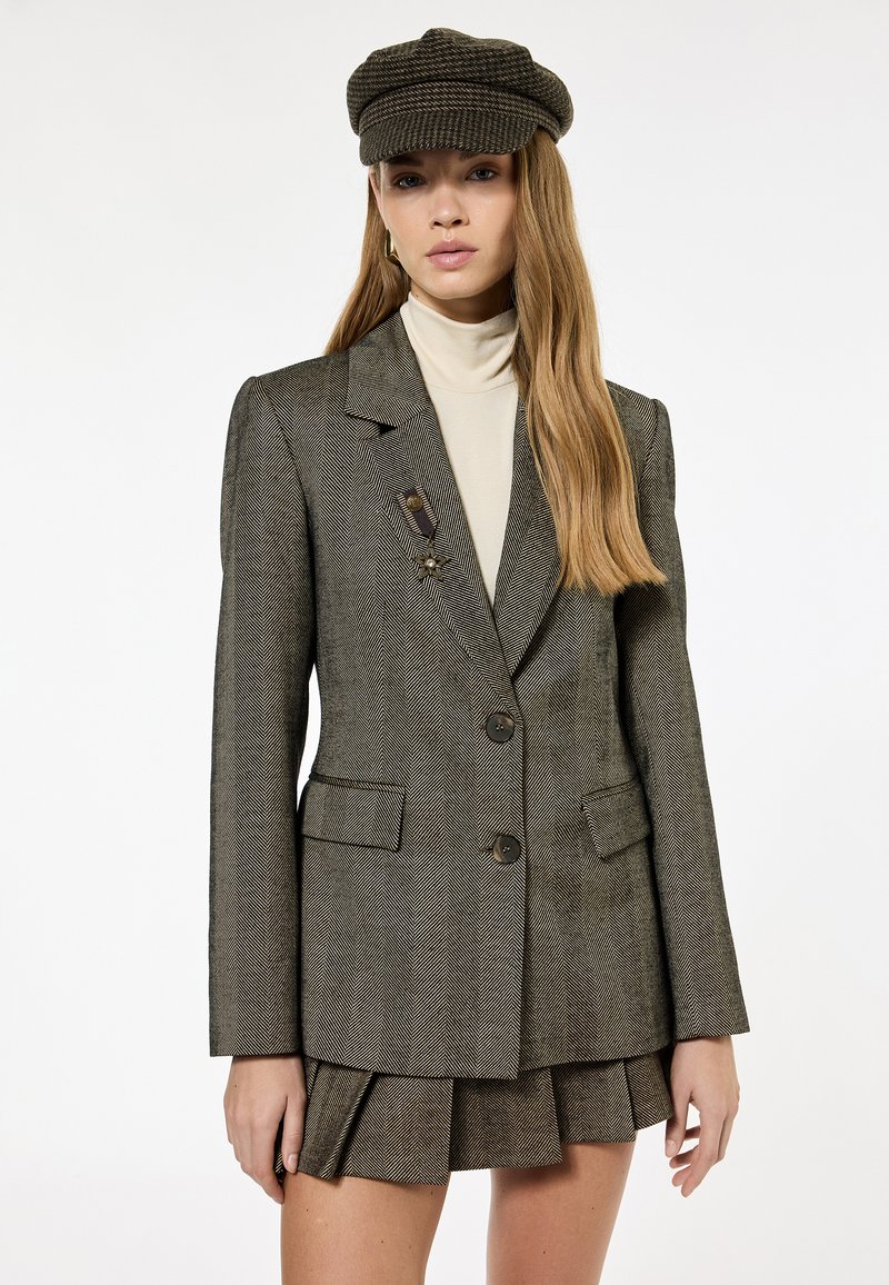 Femme portant un blazer marron à chevrons avec boutons et broche, jupe mini plissée assortie, col roulé beige et casquette gavroche marron.