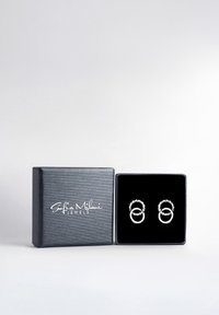 Silberohrringe mit ineinander verschlungenen, kreisförmigen Designs, präsentiert in einem schwarzen Samt-Innenraum innerhalb einer strukturierten grauen Geschenkbox.