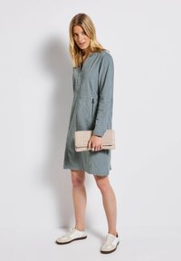 Kvinna som bär en långärmad, knälång grå klänning och vita sneakers, håller en beige vävd clutch-väska, står mot en vit bakgrund.