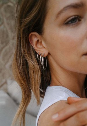Junge Frau mit hellbraunen Haaren, die drei silberne Creolen an ihrem linken Ohr trägt, gekleidet in einem weißen ärmellosen Oberteil.