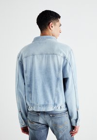 Chaqueta de denim azul claro con un corte relajado, costuras visibles, detalles de botones y un cuello, mostrada desde la parte trasera.