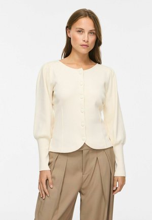 Femme aux cheveux châtain clair portant un cardigan crème boutonné et un pantalon beige plissé taille haute sur fond blanc.