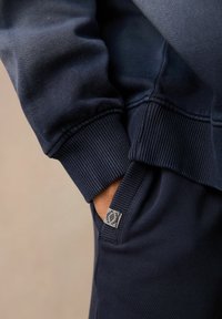 Felpa blu navy con polsini a coste e tasca laterale. La texture del tessuto è liscia, presenta un piccolo etichetta con logo sulla tasca.