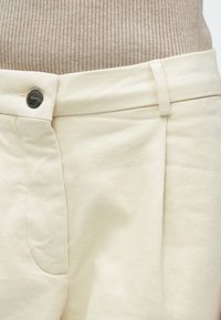 Svetlobež hlače iz tkanine z rahlo teksturirano površino, z bočnim žepom, ravnim sprednjim delom in črnim gumbom za zapenjanje v pasu.