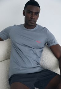 T-shirt cinza de mangas curtas com o logótipo vermelho "HUGO" no peito, combinado com calções escuros. Textura suave, design simples, silhueta ajustada.