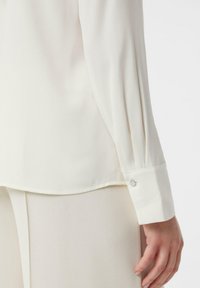 Biała, lekka bluzka z długim rękawem, z plisowanym wzorem na ramieniu i guzikami przy mankietach. Gładka tekstura.