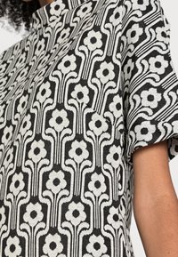Schwarz-weiß gemusterter Stoff mit floralen Motiven und vertikalen Streifen; mit Stehkragen und kurzen Ärmeln. Texturierte Webdetails sind sichtbar.