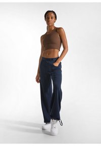 Bruine cropped tanktop gecombineerd met marineblauwe wijde pantalons met een elastische tailleband. Het model draagt witte sneakers en staat tegen een lichte achtergrond.