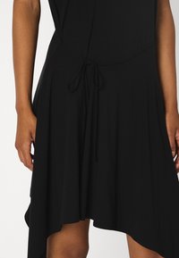 Schwarzes Kleid mit fließender Silhouette, aus leichtem Stoff gefertigt. Es hat eine Bindung in der Taille und einen asymmetrischen Saum.