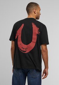 Camiseta de algodón negra con un gran diseño de pinceladas rojas en la parte posterior, mangas cortas y cuello redondo. Combinada con jeans azules.