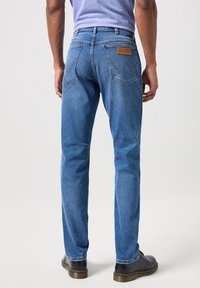 Hombre con jeans azules Wrangler y zapatos negros, de pie con la espalda hacia la cámara frente a un fondo claro y liso.