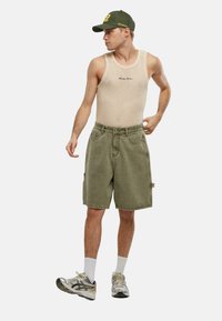 Beige nettetanktopp med brodert logo, kombinert med olivengrønne cargo-shortser. Modellen har på seg hvite crew-sokker og sølvfargede joggesko.