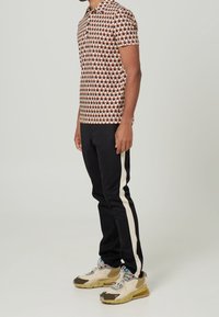 Polo à manches courtes avec un motif géométrique rouge et crème, associé à un pantalon noir au liseré crème et des baskets de sport beiges.