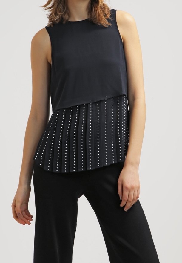 Femme portant un haut noir sans manches avec un ourlet asymétrique plissé clouté et un pantalon noir sur fond uni.