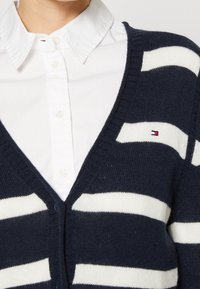 Cardigan blu navy con righe orizzontali bianche, design a V, bordi a costine e chiusura con bottoni. Indossato sopra una camicia bianca con colletto.