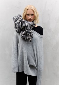 Pull gris oversize en maille douce, associé à un top noir. Une écharpe noire et grise à motifs est drapée autour du cou, avec des bords à franges.