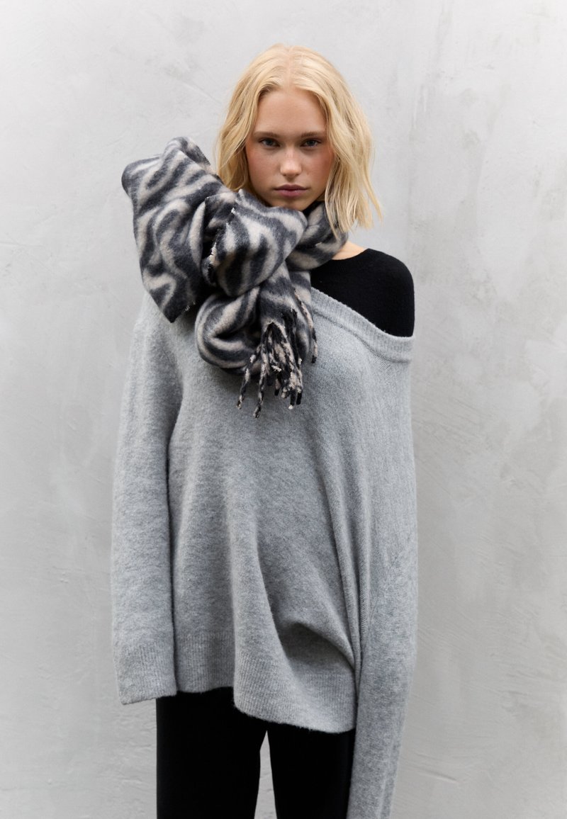 Pull gris oversize en maille douce, associé à un top noir. Une écharpe noire et grise à motifs est drapée autour du cou, avec des bords à franges.