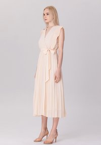 Robe plissée pêche pâle avec un décolleté en V profond, des manches volantes et une ceinture à nouer à la taille. Tissu lisse avec une texture fluide.