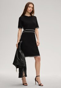 Μαύρο φόρεμα με κοντά μανίκια, λευκή επένδυση, εφαρμοστό σχέδιο, μήκος μέχρι το γόνατο. Το μοντέλο κρατά δερμάτινο μαύρο μπουφάν και φορά μαύρα σανδάλια με τακούνι.