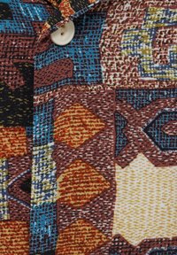 Chemise à motifs présentant un mélange de tons terreux en formes géométriques. Le tissu est texturé avec un détail bouton au col.