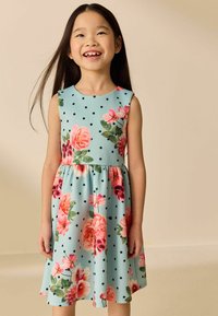 Fille souriante aux longs cheveux foncés portant une robe bleu sans manches décorée de fleurs roses et de pois noirs sur un fond beige.
