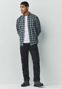 Young man standing with hands behind back, wearing gray checkered shirt over white t-shirt, black pants, and black sneakers.