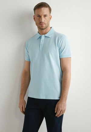 Mężczyzna z krótkimi włosami, ubrany w jasnoniebieską koszulkę polo z krótkim rękawem i ciemne spodnie, stojący na tle jednolitej białej ściany.