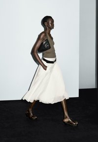 Jupe longue blanche, débardeur marron, sac noir. Sandales à plateforme marron avec des clous. Modèle marchant devant un fond neutre.