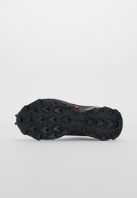 Semelle d'une chaussure de trail running noire avec des crampons profonds en forme de flèche agressive et un petit détail rouge près du centre.