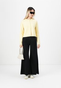 Cardigan en maille côtelée jaune, pantalons larges noirs, baskets blanches et un petit sac à main blanc. Les accessoires comprennent des lunettes de soleil sombres.