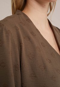 Brūns audums ar teksturētu rakstu un v-veida dizainu, ar smalkiem tonāliem akcentiem. Gluda tekstūra ar plūstošu, mīkstu audumu.