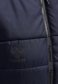 Chaqueta de plumas azul marino con un patrón de cuadrícula cosido, que presenta un logotipo tonal y un ribete negro en el zipper con el nombre de la marca "hummel".