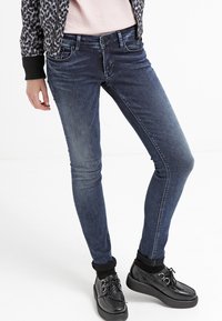 Jeans ajustados de denim de lavado oscuro con una textura suave, que presentan un diseño clásico de cinco bolsillos y un ligero desgaste. Combinados con zapatillas negras.