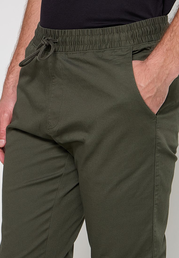 Jogging en jersey à taille élastique en olive, présentant des poches latérales, une texture lisse, un cordon de serrage ajustable et un design décontracté.