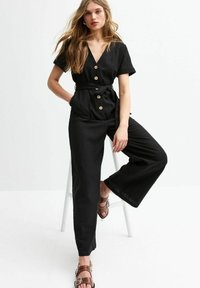 Combinaison noire à manches courtes, décolleté en V, fermeture à boutons sur le devant, taille nouée, design à jambes larges et tissu texturé. Associée à des sandales brunes.