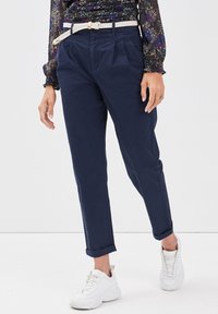 Femme portant un pantalon fuselé bleu marine avec une ceinture tressée blanche, un chemisier à manches longues fleuri et des baskets blanches, debout sur un sol blanc.