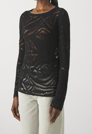 Haut en dentelle noir à manches longues avec des motifs abstraits porté avec un pantalon beige clair sur une personne devant un fond clair.