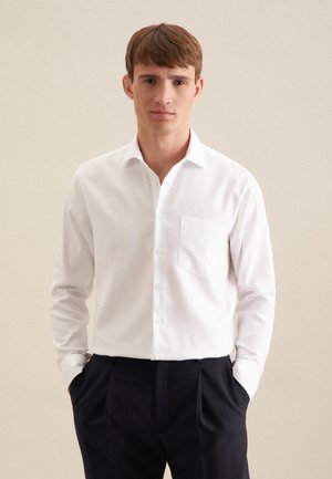 Jeune homme aux cheveux courts bruns portant une chemise blanche boutonnée et un pantalon sombre, les mains dans les poches, devant un fond uni.