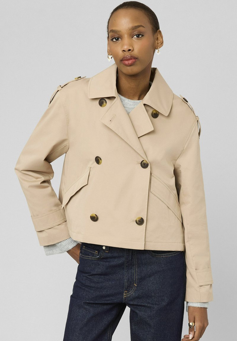 Femme portant un trench-coat court beige à double boutonnage, associé à un jean bleu foncé et un pull gris en dessous.