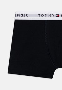 Melni kokvilnas bokseršorti ar baltu elastīgo jostu, uz kuras melnā krāsā ir uzdrukāts "TOMMY HILFIGER" logo. Gluda tekstūra, klasiskā piegriezuma.