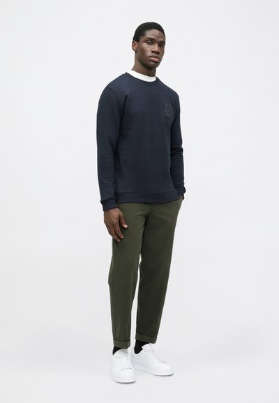 Marineblå bomulds sweatshirt med lange ærmer og rund hals. Matchet med olivengrønne tapered bukser og hvide sneakers. Diskret logo detalje på sweatshirten.